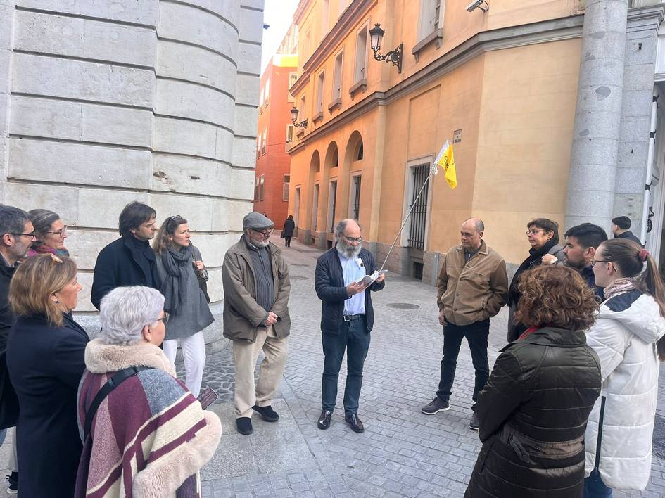 Un grupo de personas escucha atentamente a un hombre que sostiene un objeto, en una calle de una ciudad.