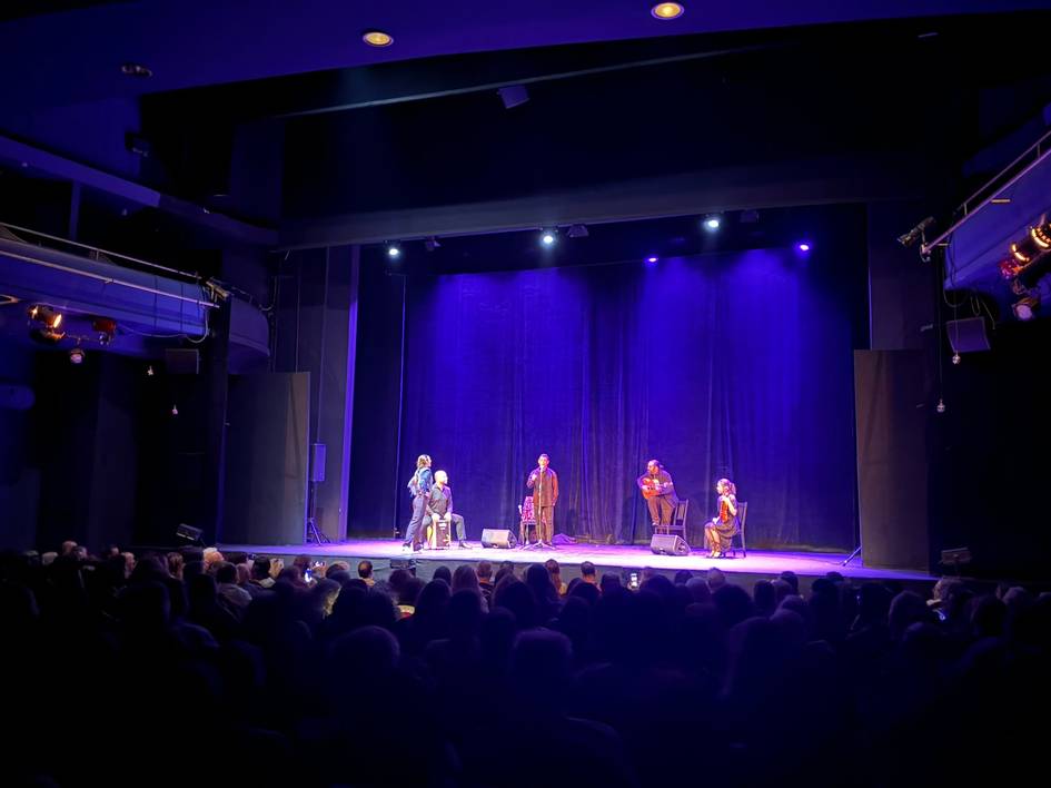 Un grupo de músicos se presenta en un escenario iluminado con luces moradas ante un público atento.
