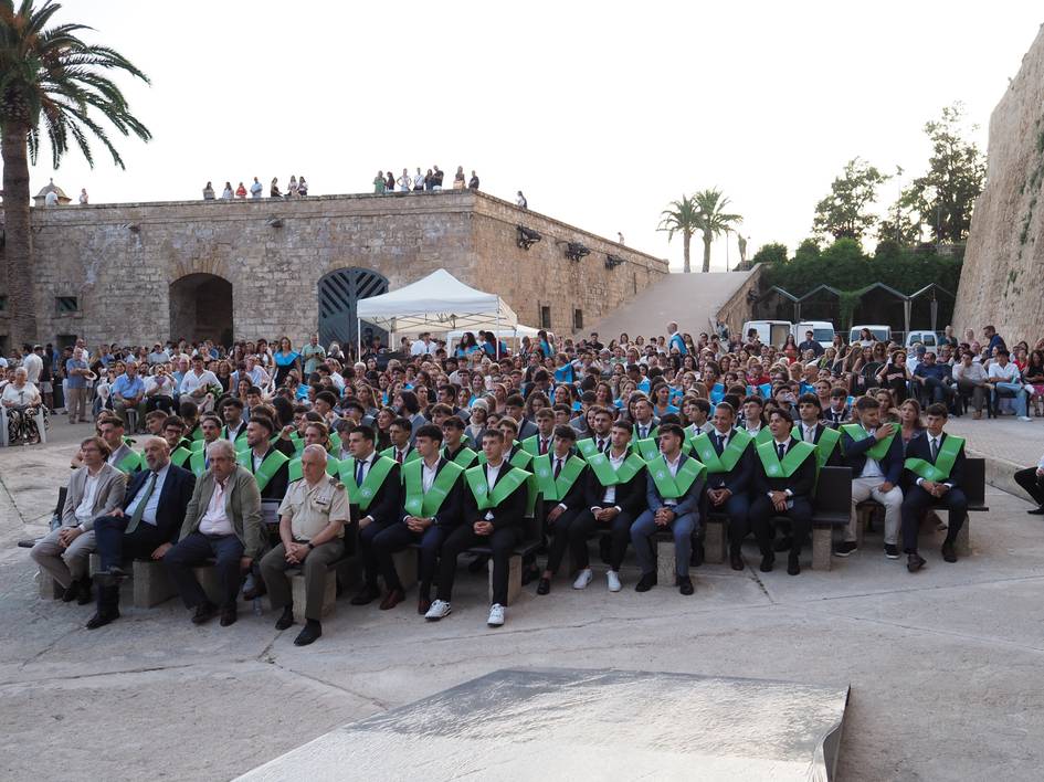 Un grupo numeroso de personas asistiendo a una ceremonia al aire libre, con una gran cantidad de graduandos en primera fila vistiendo togas con capes verdes.