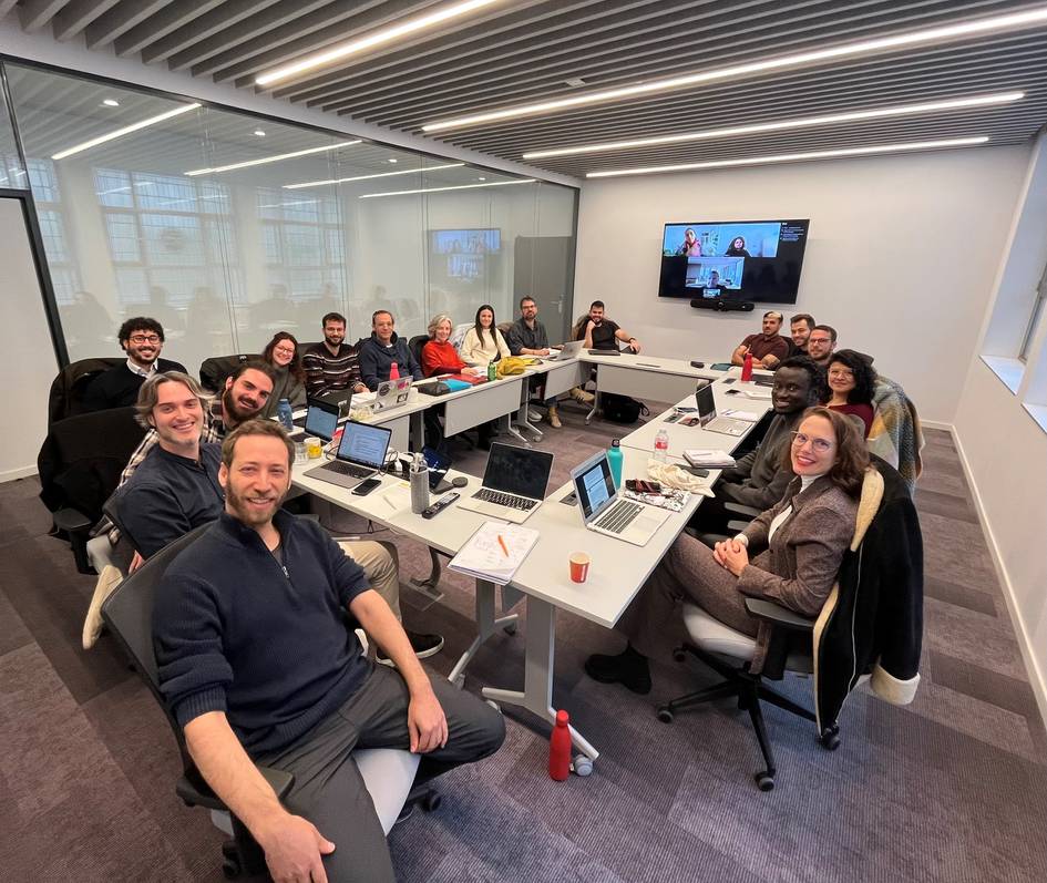 Un grupo de personas sonrientes se encuentra reunido en una sala de conferencias moderna, con computadoras portátiles y una pantalla de videoconferencia.