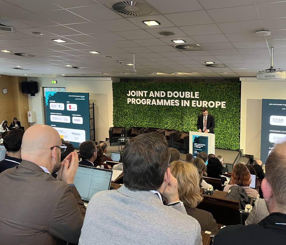 Una conferencia con un presentador hablando frente a un público atento en un entorno académico.