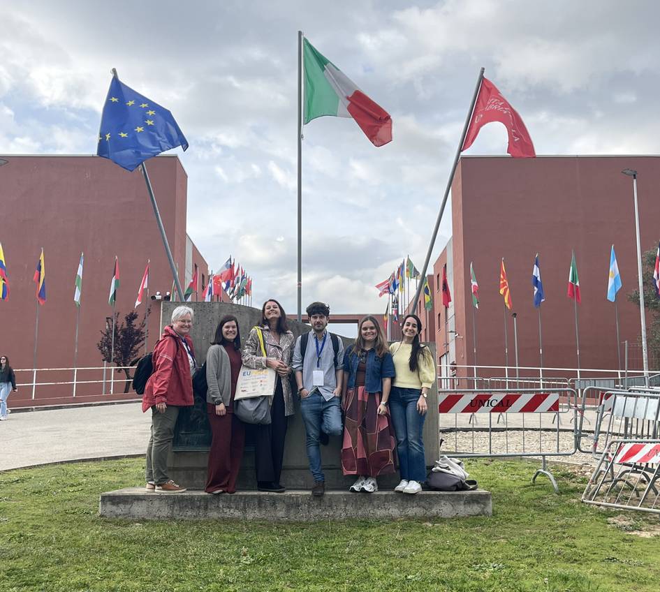Un grupo de seis personas posan frente a varias banderas en un lugar al aire libre.