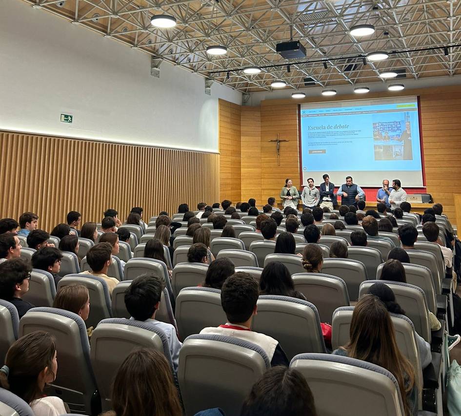 A group of people attending a presentation in a modern auditorium with panelists speaking at the front.