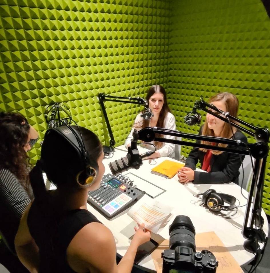 Cuatro mujeres participan en un podcast, sentadas alrededor de una mesa con micrófonos en una sala con paredes verdes insonorizadas.