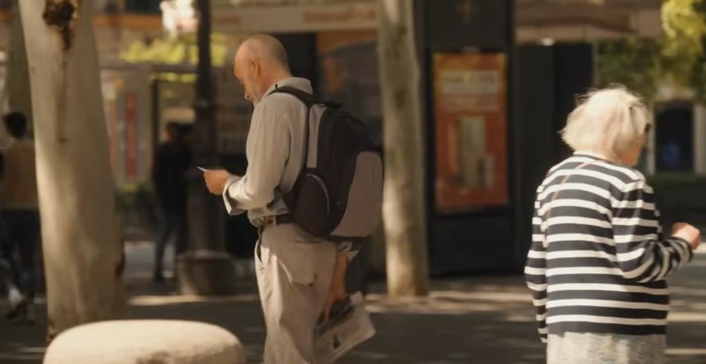 An older man and woman walking on a sunny city street, the man is using a smartphone.