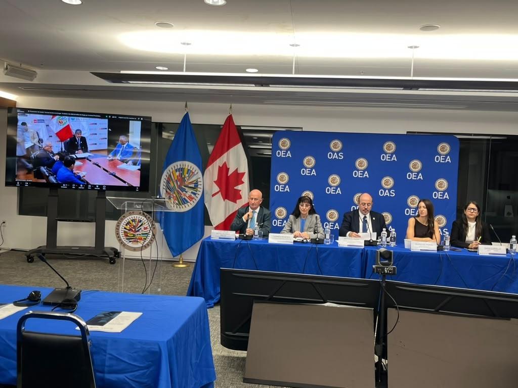La imagen muestra una conferencia de prensa con varios panelistas y banderas de Canadá y la OEA en el fondo.