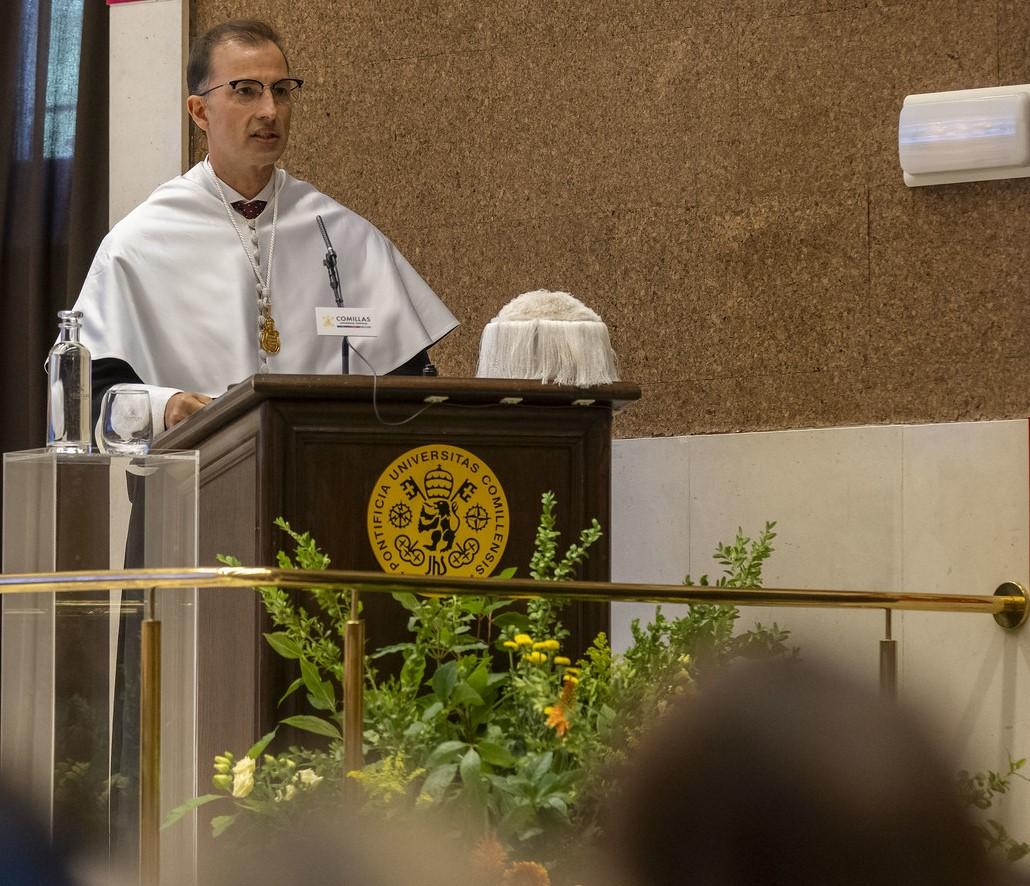 Un hombre vestido con una capa blanca y gafas habla desde un podio en un evento formal.