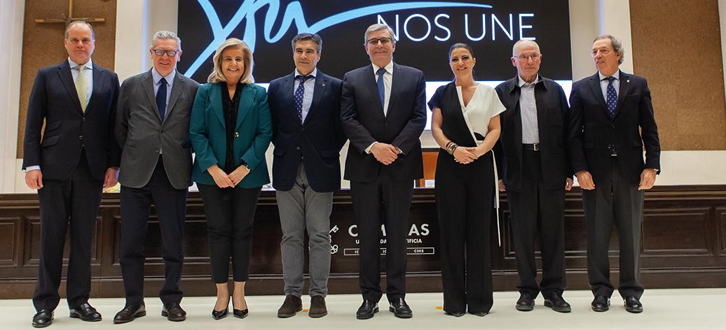 Grupo de ocho personas, cuatro hombres y cuatro mujeres, de pie en un evento formal frente a un cartel que dice 'XXI Nos Une'.