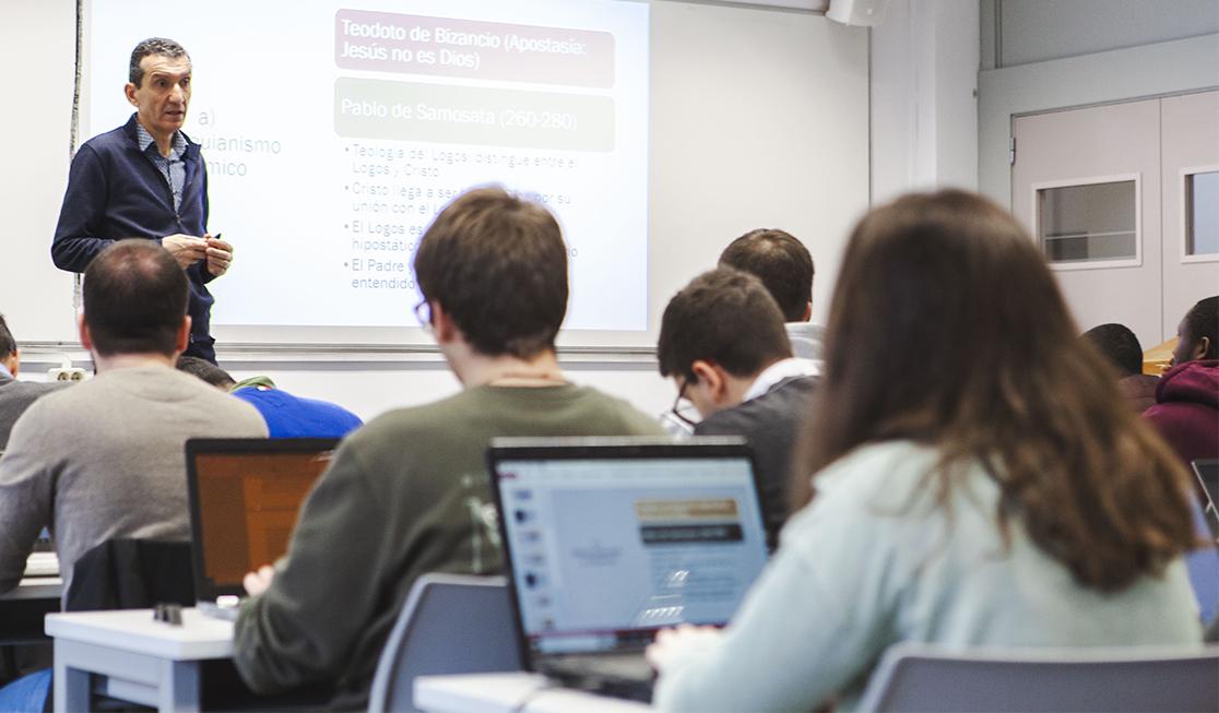 Un profesor está dando una charla en un aula mientras los estudiantes trabajan en sus computadoras portátiles.