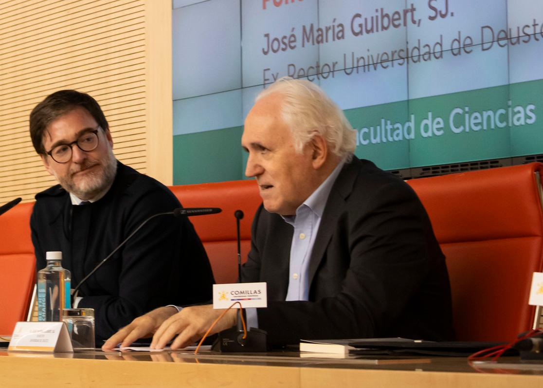 Dos hombres en una mesa durante una conferencia, uno vestido con sotana y el otro con traje y corbata.