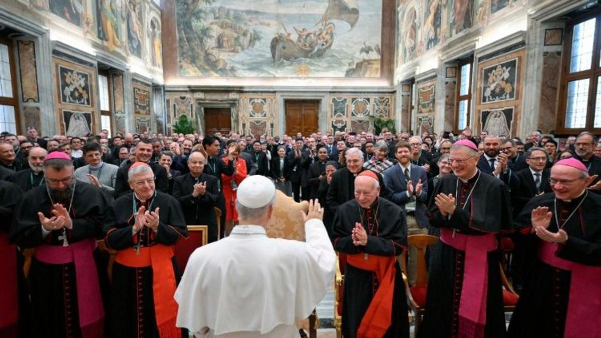 Una multitud de clérigos asisten a un evento en una sala decorada con frescos, donde un líder religioso se dirige a ellos.
