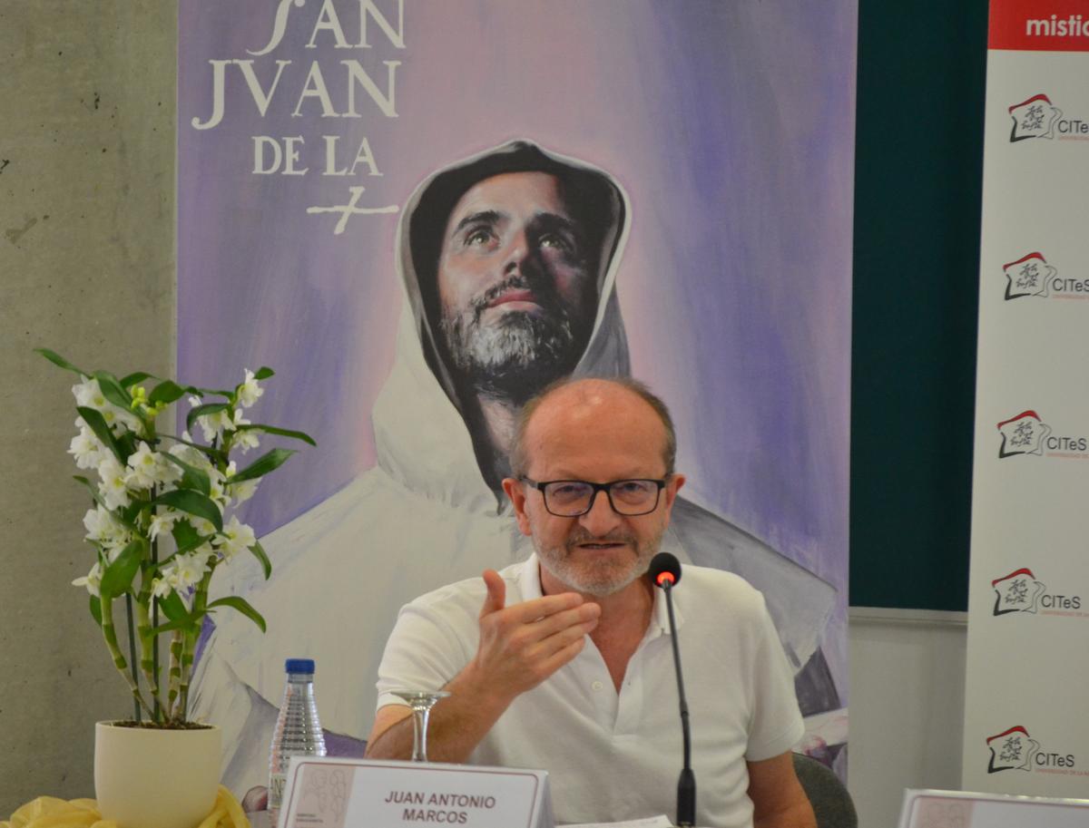 Un hombre habla frente a una mesa con un cartel de San Juan de la Cruz detrás de él.