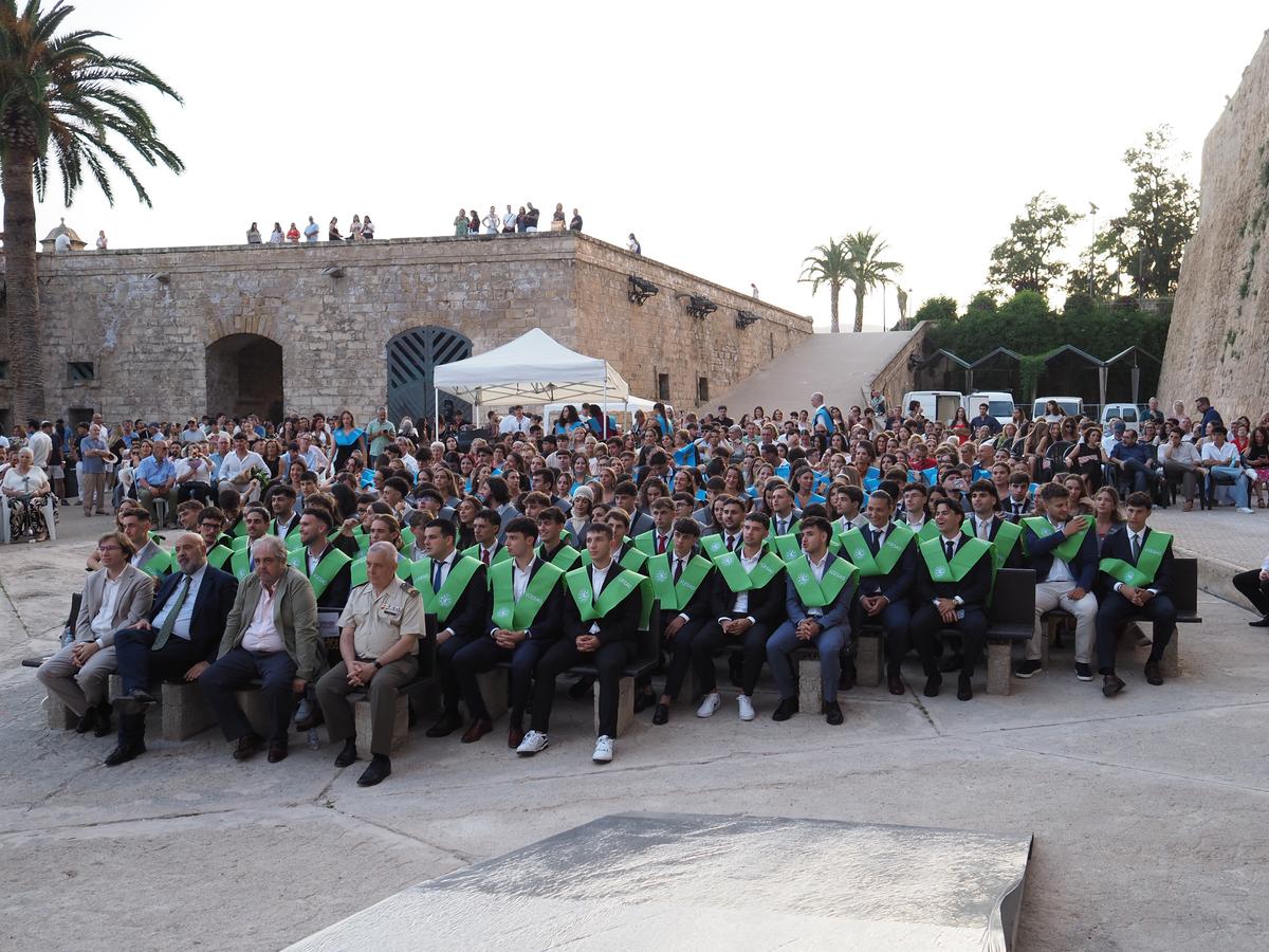 Un grupo numeroso de personas asistiendo a una ceremonia al aire libre, con una gran cantidad de graduandos en primera fila vistiendo togas con capes verdes.