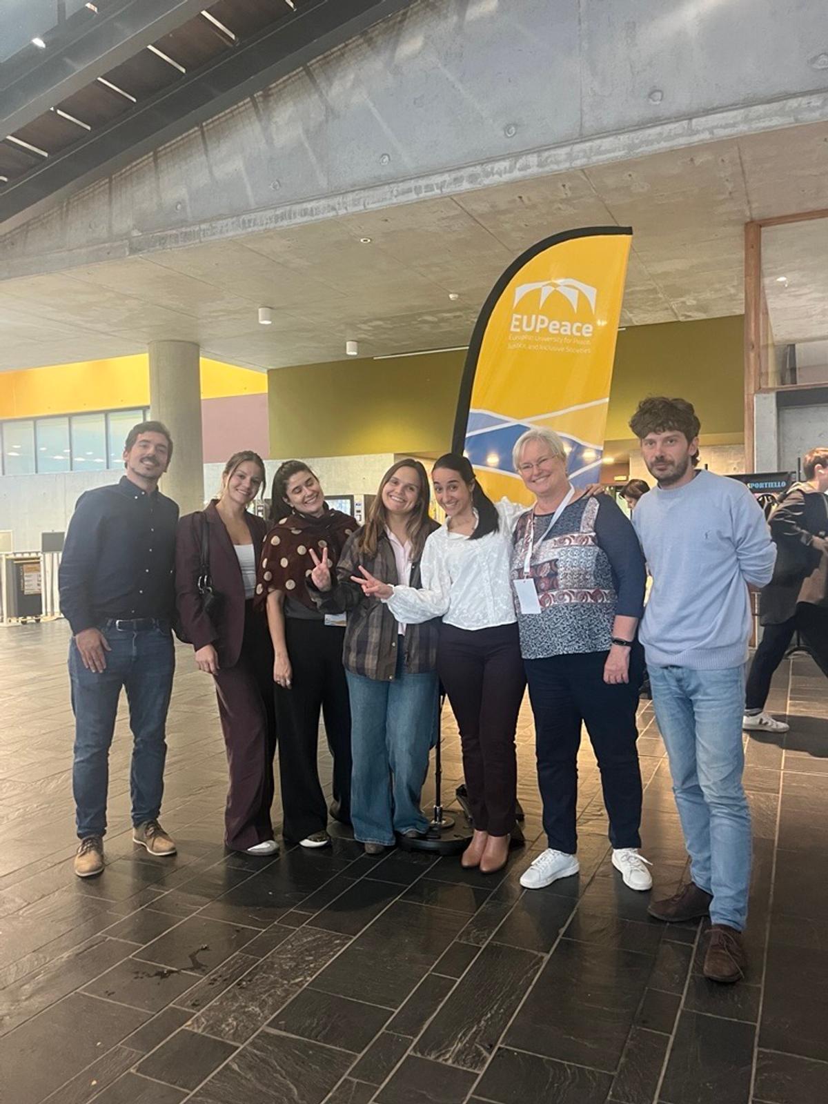 Un grupo de personas sonrientes posando juntas frente a un banner de EUPeace en un ambiente interior moderno.