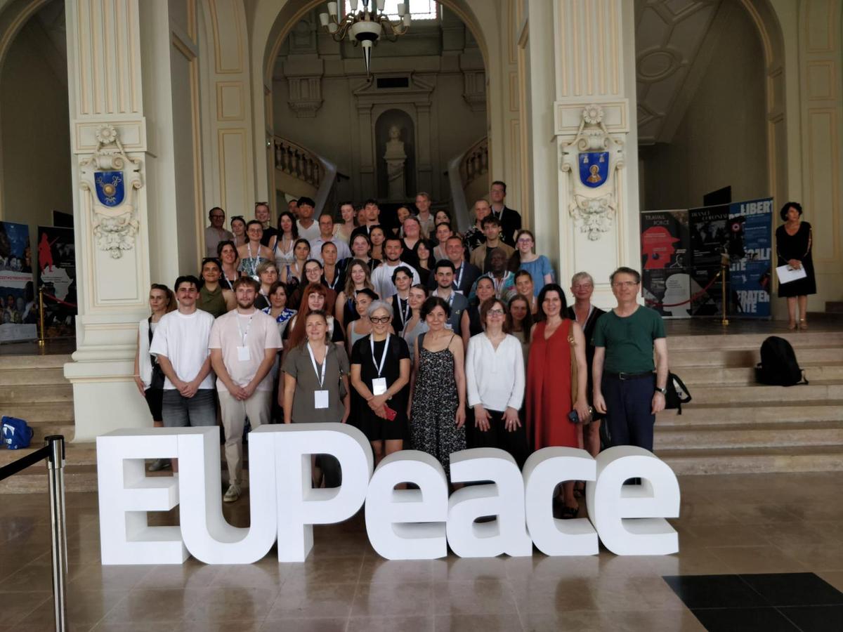 Un grupo diverso de personas posando frente a un cartel que dice 'EUPeace' en un edificio elegante.