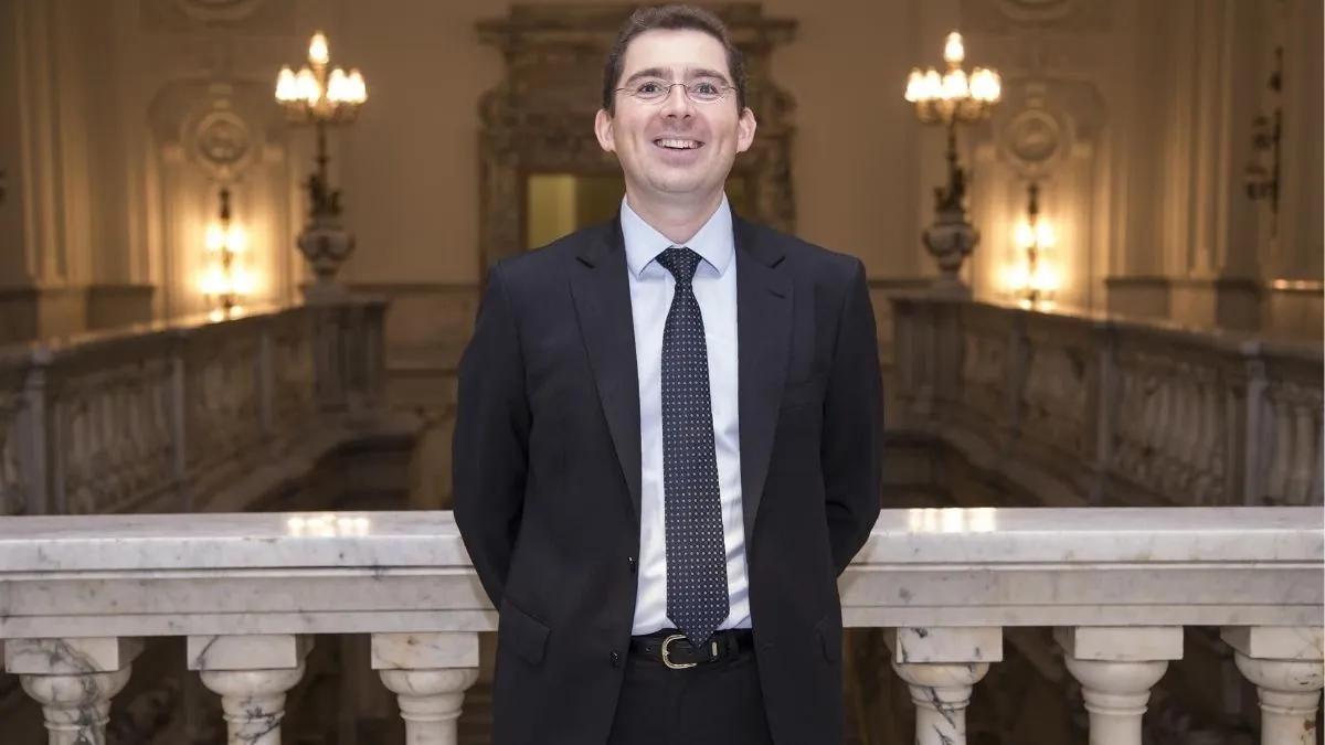 Un hombre sonriente vestido de traje se encuentra de pie en un vestíbulo elegante con decoración clásica.