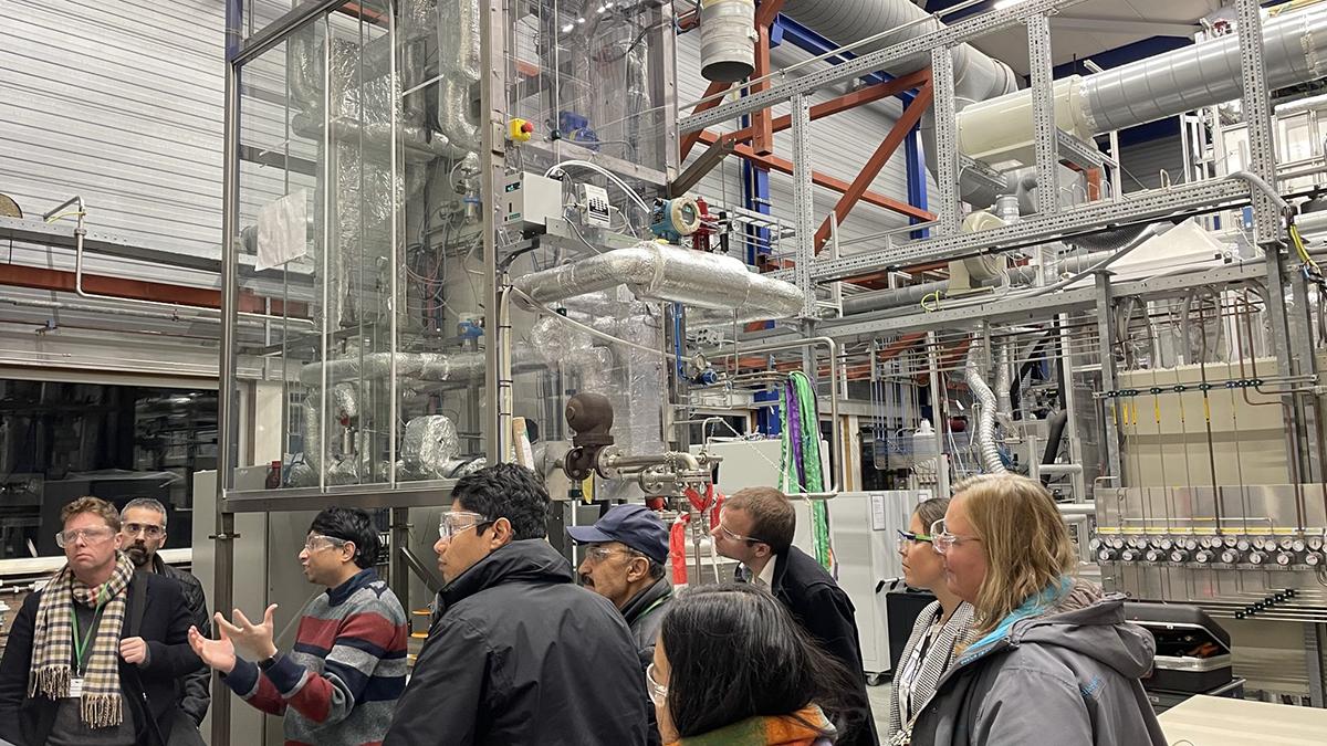 Un grupo de personas recibe una explicación en un complejo industrial con grandes tuberías y maquinaria.