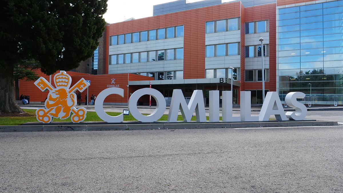 La imagen muestra el letrero de la Universidad Pontificia Comillas, con un edificio moderno al fondo.