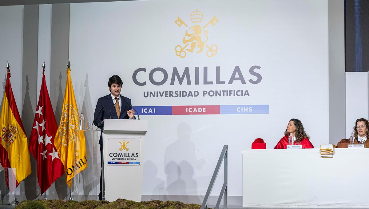 Un hombre habla en el escenario de un evento en la Universidad Pontificia Comillas, rodeado de banderas y un panel informativo.