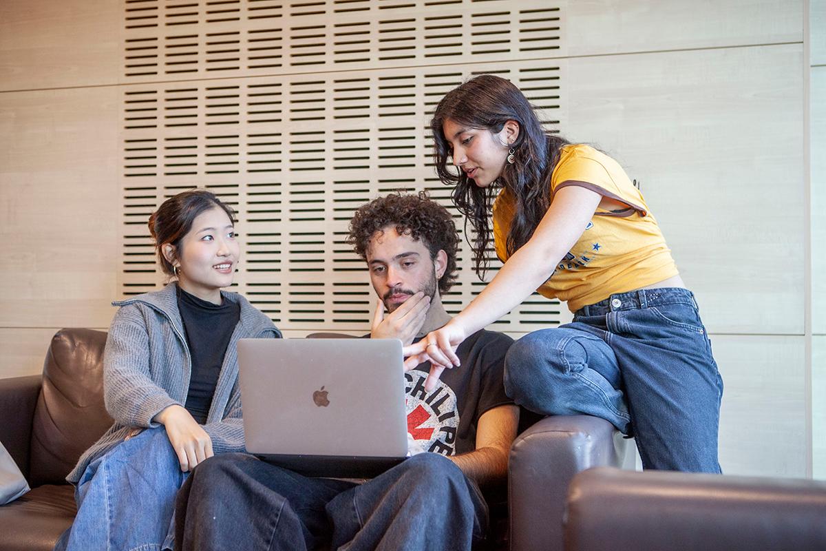 Tres personas están sentadas en un sofá, mirando una computadora portátil y conversando entre ellas.