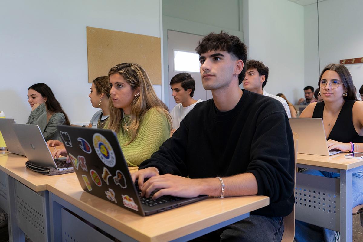 Un grupo de estudiantes está sentado en un aula con laptops, prestando atención a una clase.