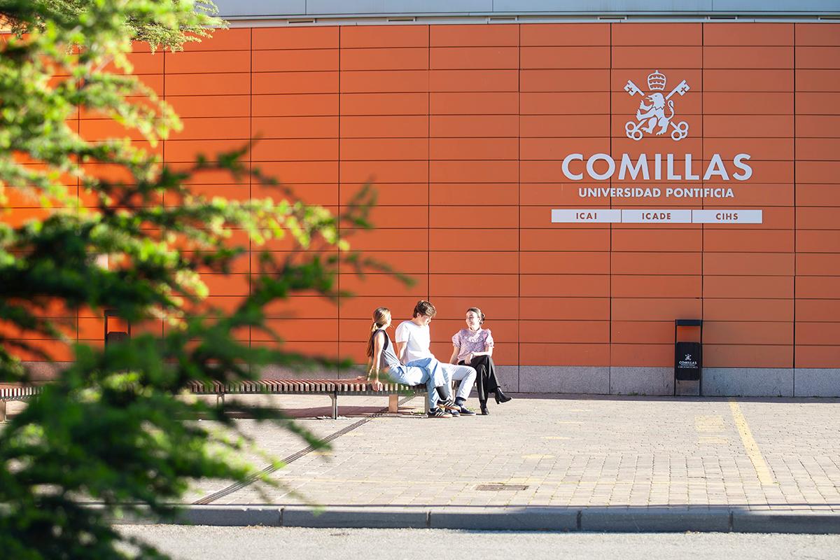 Un grupo de estudiantes se sienta en un banco frente a un edificio de la Universidad Pontificia Comillas.