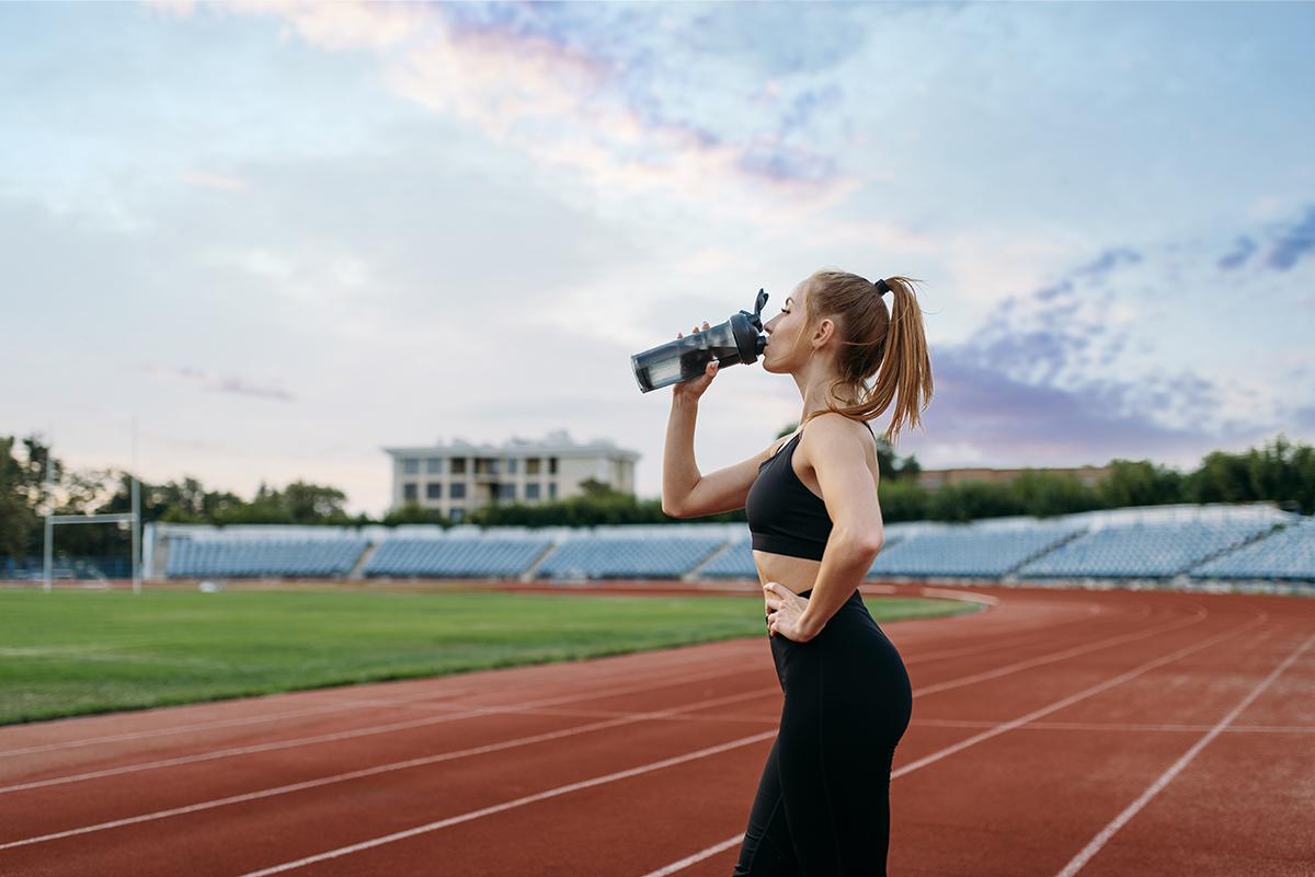 Mujer bebiendo agua en una pista de atletismo al atardecer.