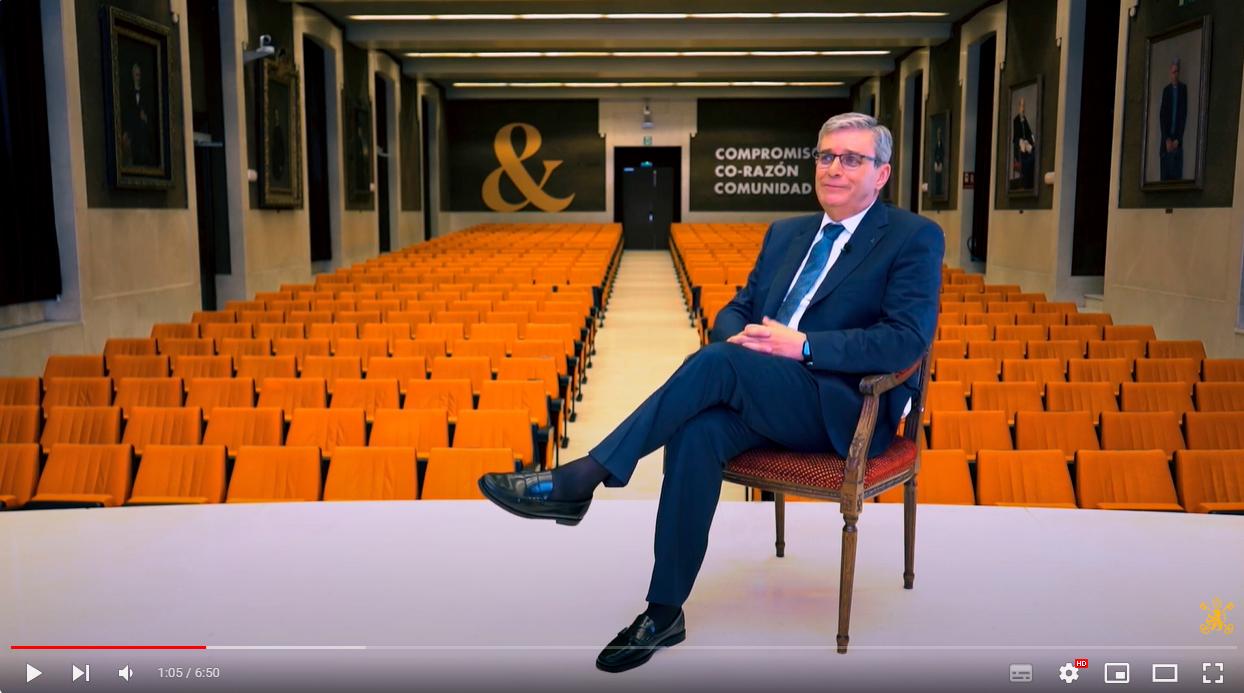 A man in a suit seated on a chair in an empty auditorium with orange seats and a screen displaying text behind him.