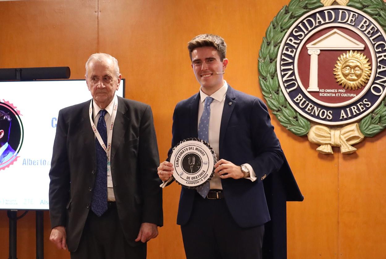 Two men, one elderly and one young, holding a commemorative plate, standing in a room with academic insignias