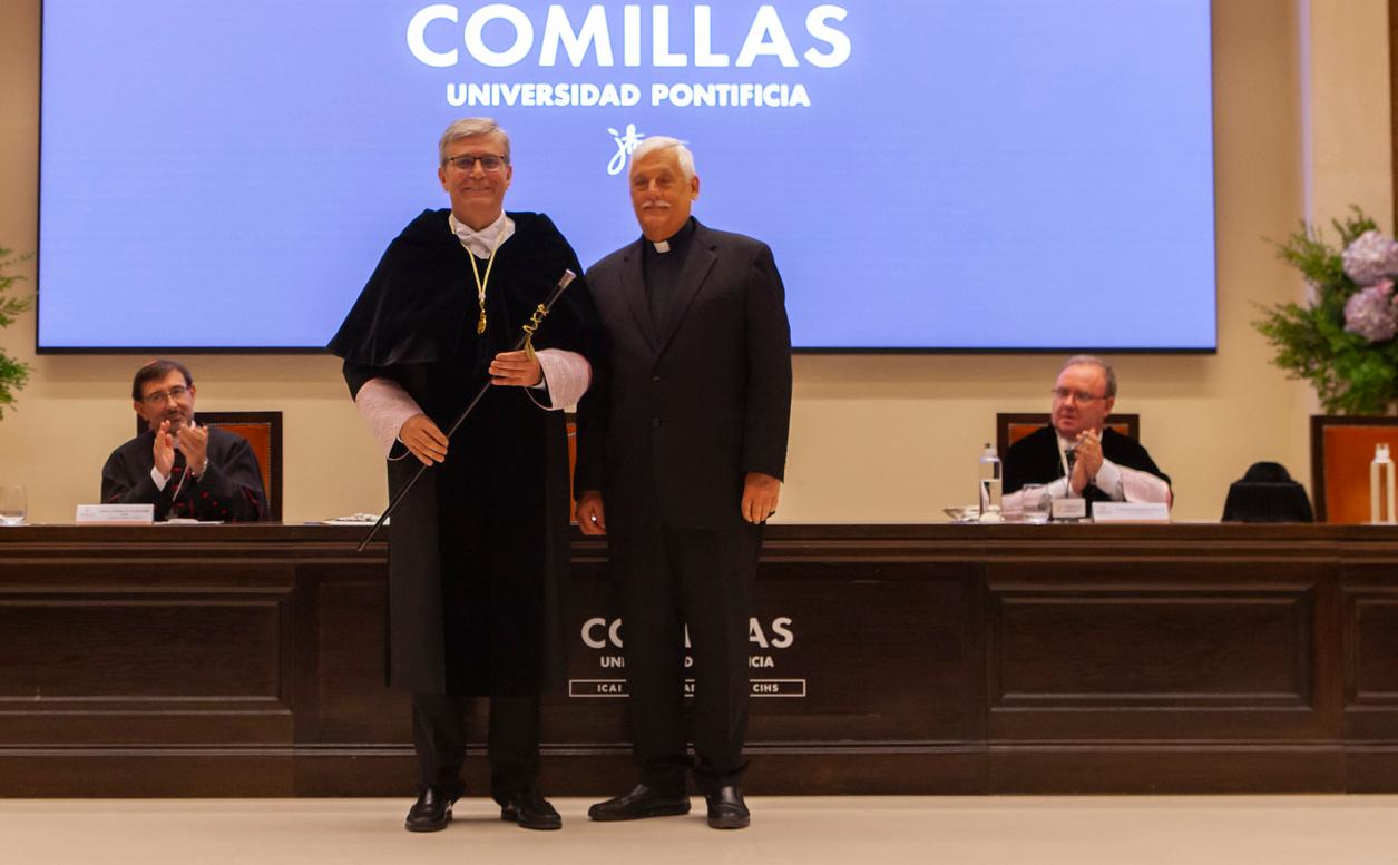 Two men in academic attire posing with a staff, standing in front of a sign for Comillas Pontifical University.