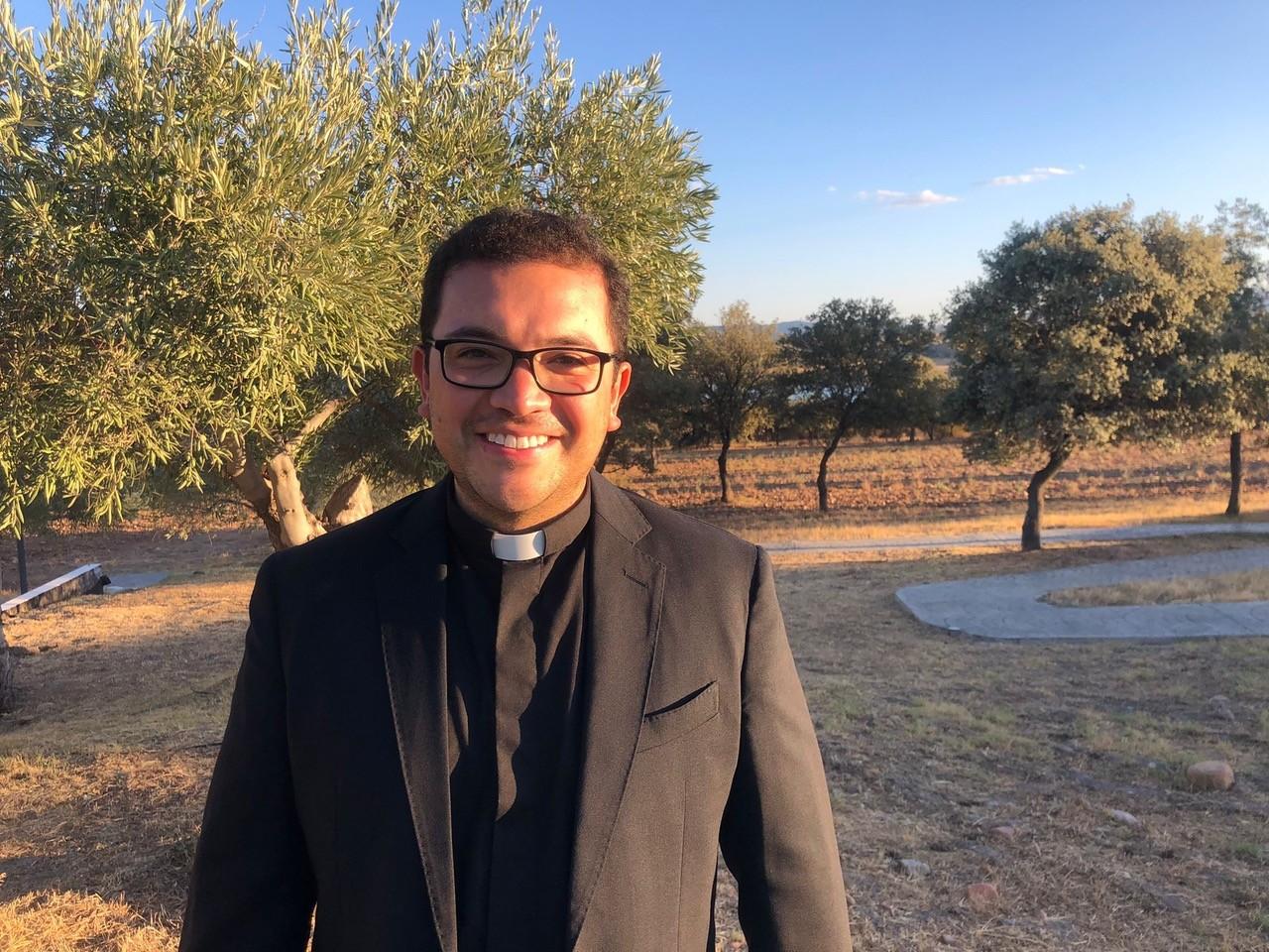 Un sacerdote sonriente en un entorno natural con árboles y luz solar al atardecer.