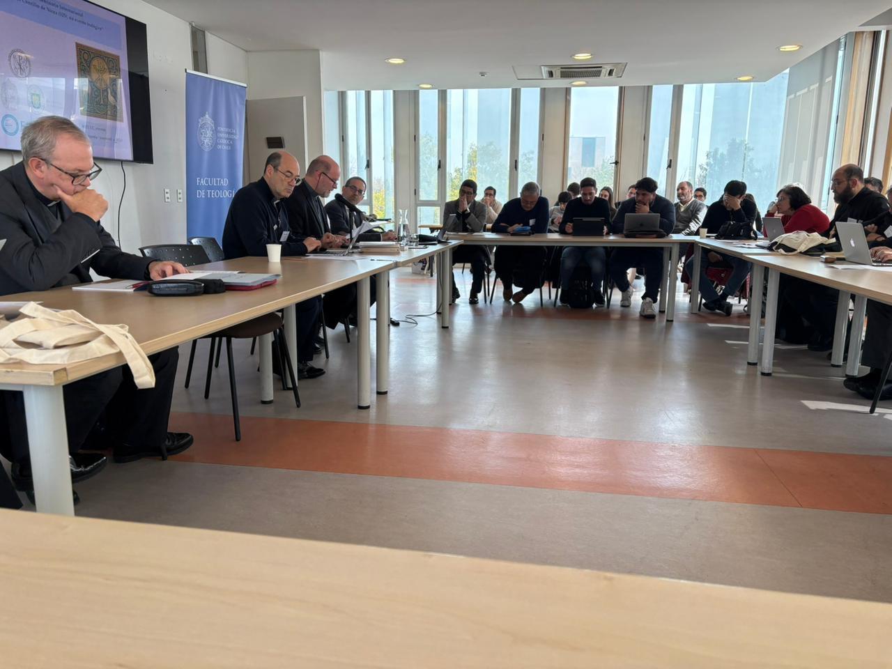 Reunión de un grupo de personas sentadas en una mesa, prestando atención a un orador al frente en un ambiente luminoso.