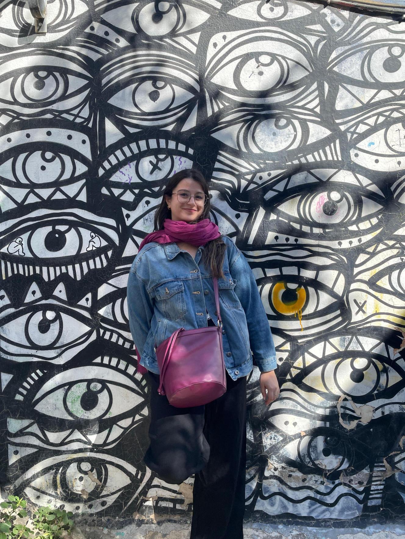 Una mujer posa frente a un mural de grandes ojos en una pared.