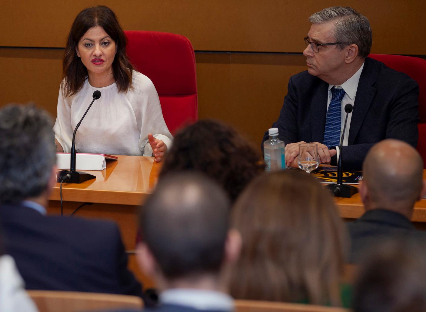 A woman speaks animatedly at a conference table while a man listens, surrounded by an attentive audience.