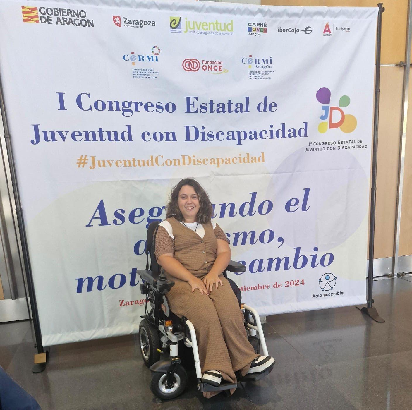 Una mujer sonriendo en una silla de ruedas delante de un banner del I Congreso Estatal de Juventud con Discapacidad.