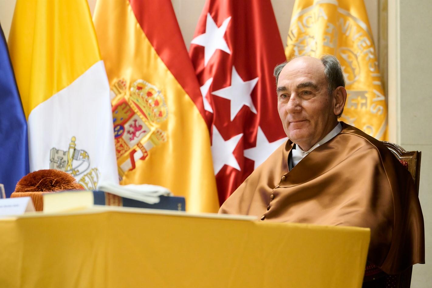 An elderly man in academic robes sits at a desk with national flags in the background.