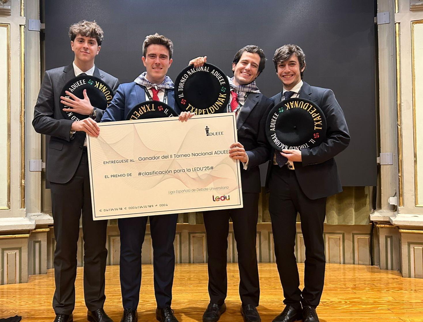 Four young men in suits holding a large ceremonial check and awards inside a formal venue.