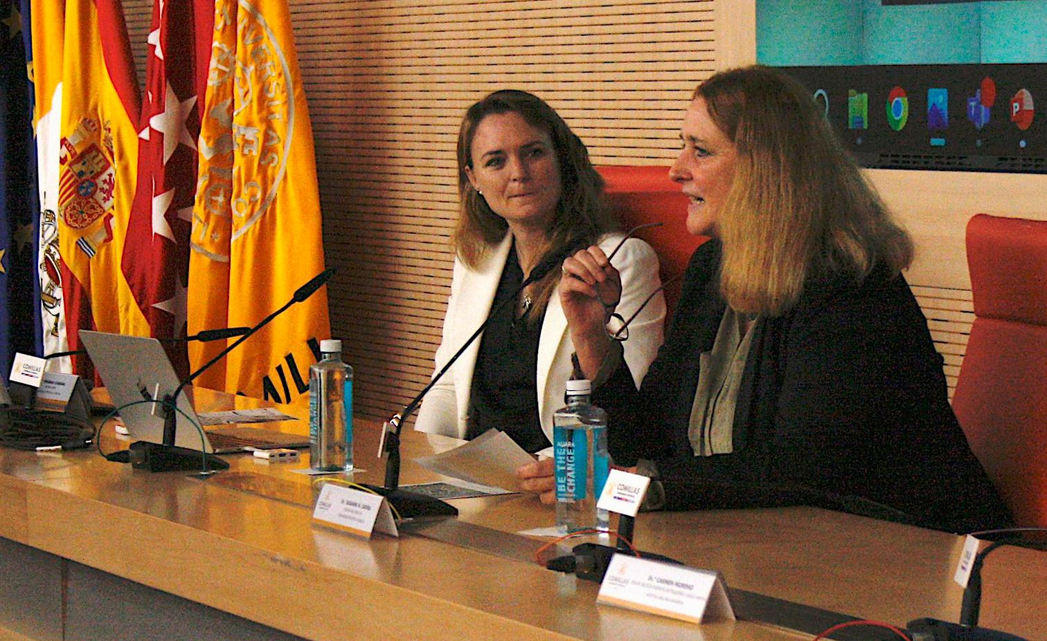 Two women are engaged in a discussion during a conference, seated at a table with microphones and water bottles, in a room with flags.