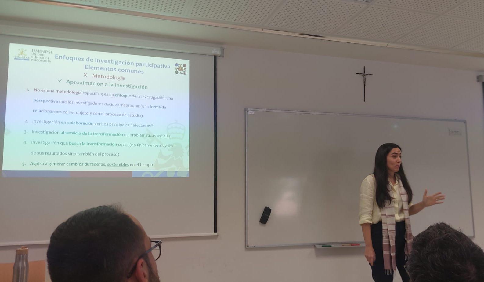 A woman presenting a scientific topic in a classroom with a PowerPoint slide in the background.