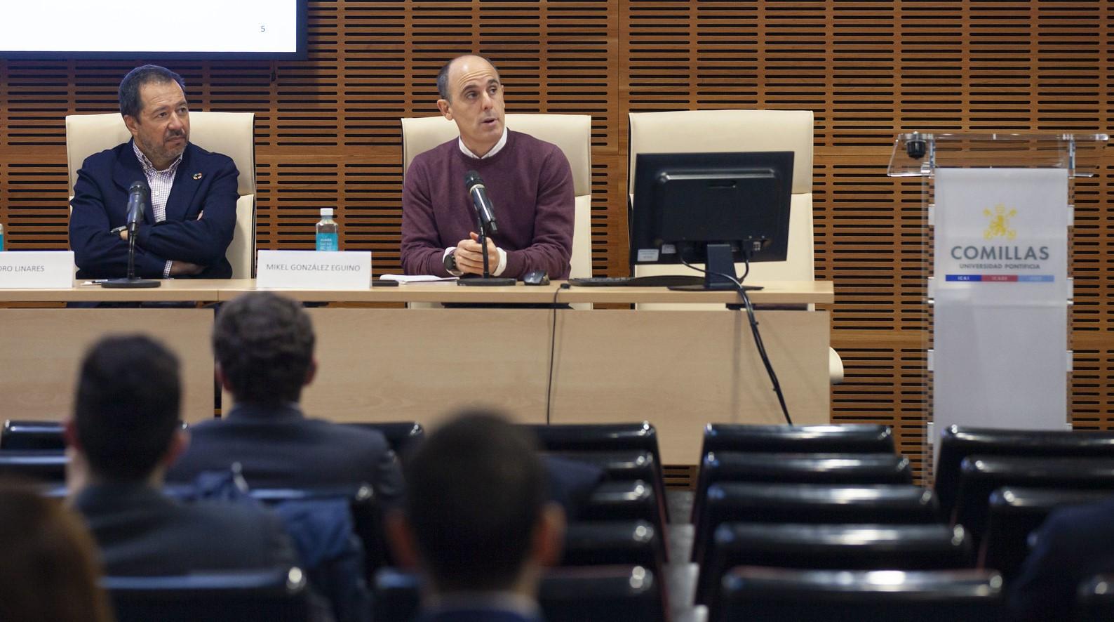 Dos hombres están sentados en un panel frente a una audiencia en una sala de conferencias.