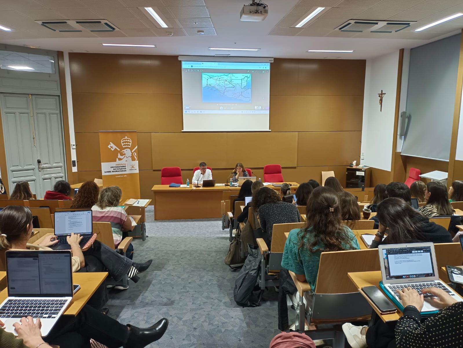 A group of students attentively sitting in a university lecture hall while a presenter discusses a topic at the front.