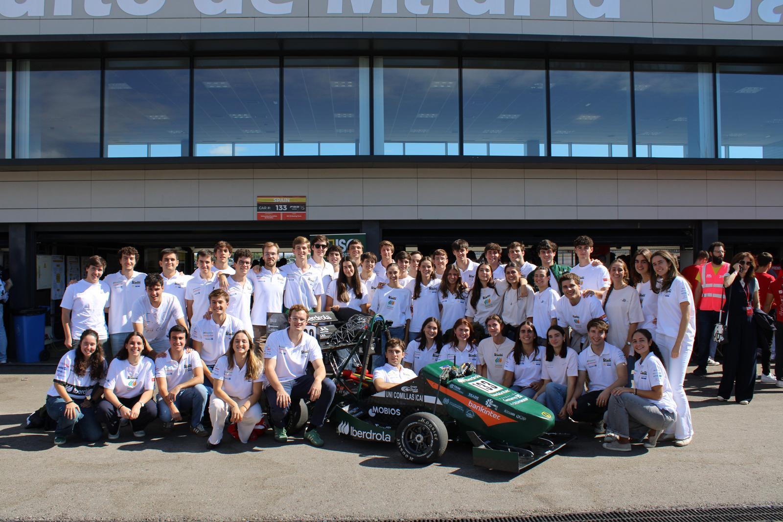 Un grupo de estudiantes sonrientes posa en frente de un coche de carreras en un evento relacionado con la ingeniería.