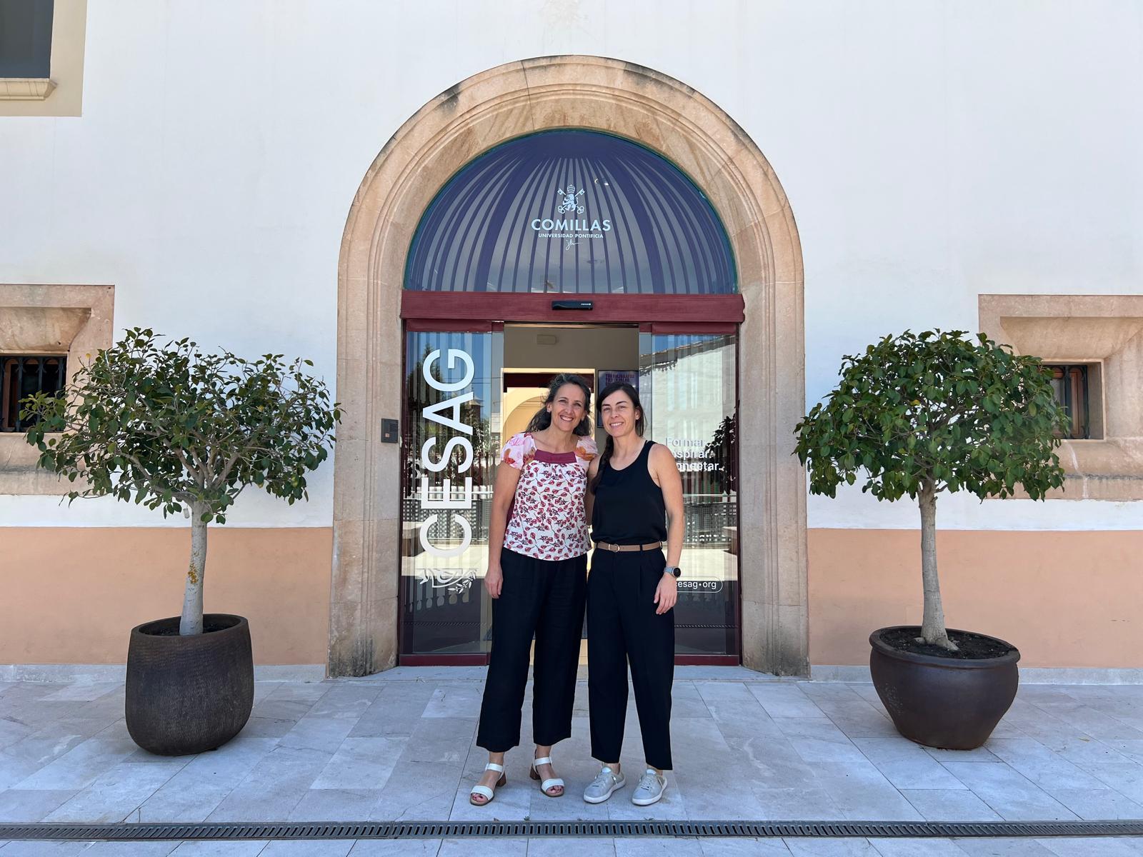 Dos mujeres posan sonrientes frente a un edificio con plantas a los lados de la puerta de entrada.