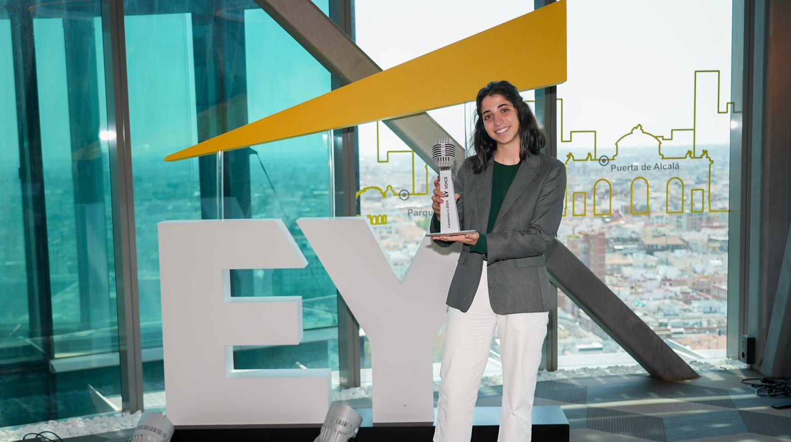 A woman stands smiling, holding an award next to a large ‘EY’ sign with a cityscape in the background.