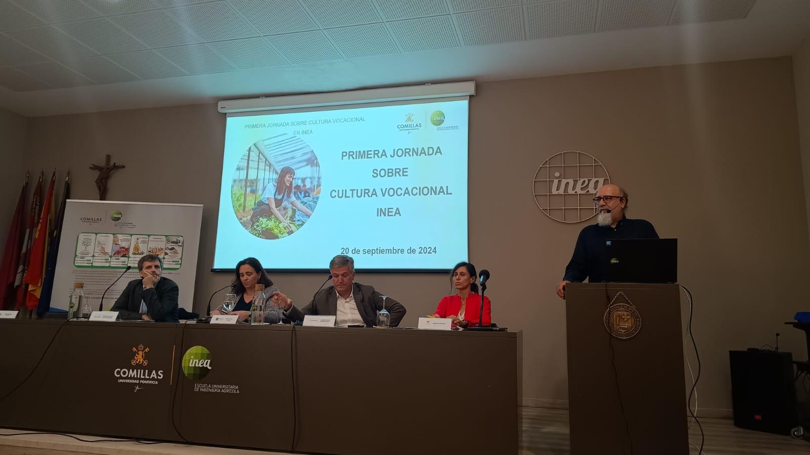 A man is speaking at a podium during a conference event, with four other participants seated at a table with a banner of 'Comillas Pontifical University' in the background.