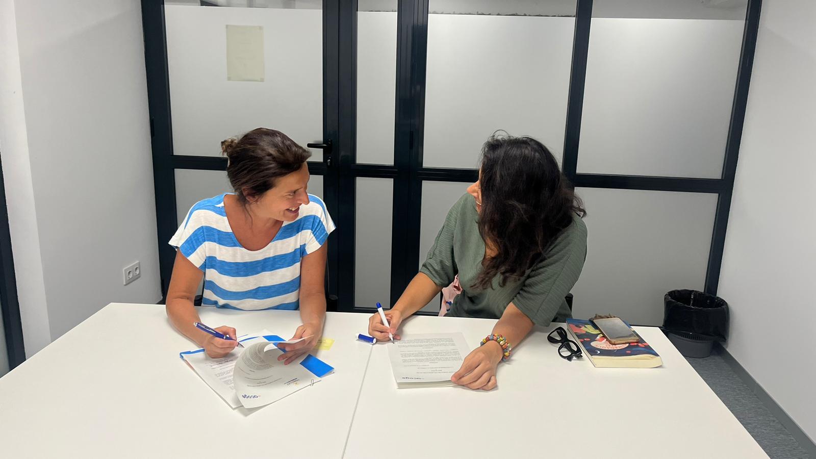 Two women are sitting at a table in an office, discussing documents.