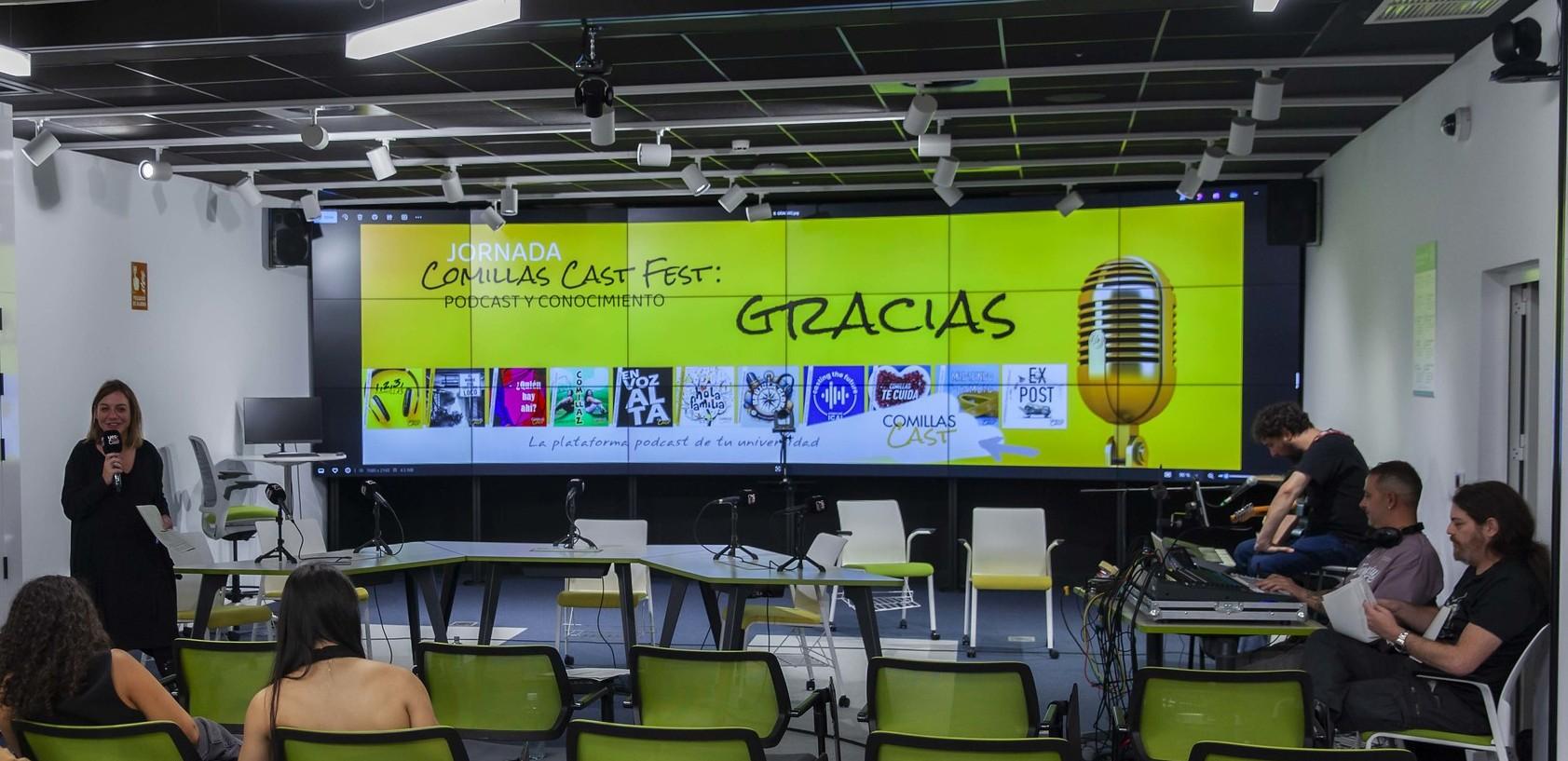 Una mujer está hablando frente a un micrófono en una sala con varias personas sentadas y una pantalla grande al fondo que dice 'GRACIAS' junto a varios logos.