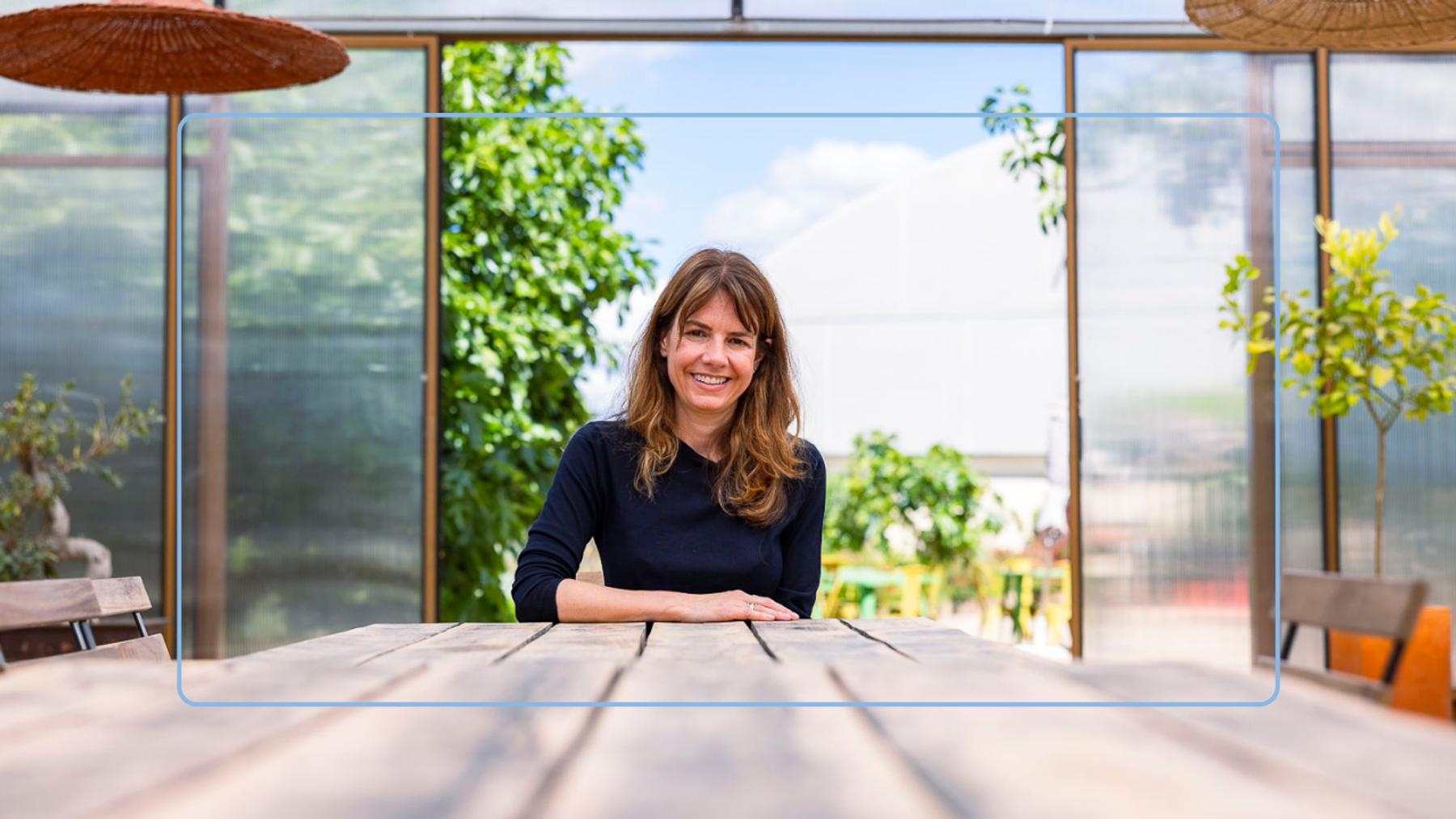 Una mujer sonriente se sienta en una mesa de madera en un entorno luminoso y natural.
