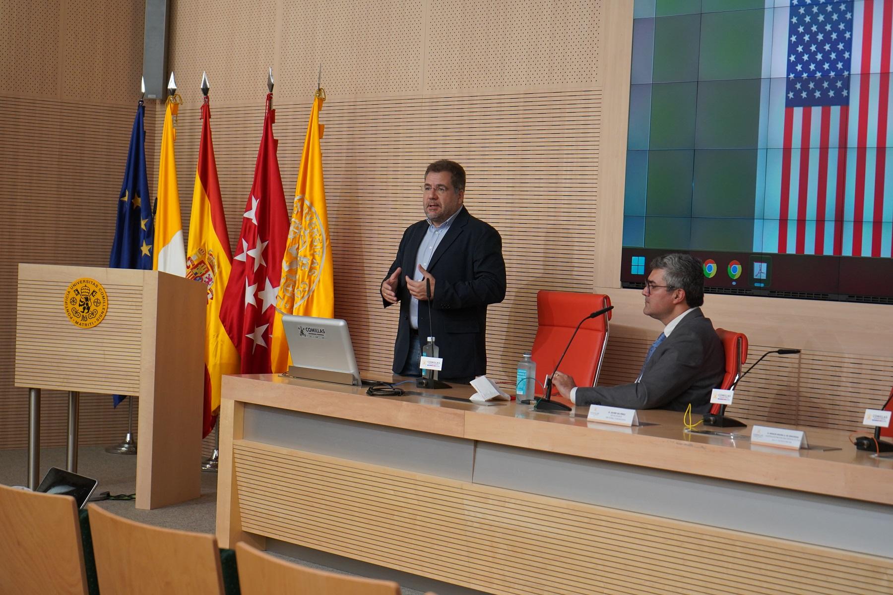 A man is speaking at a podium in a conference room with flags of Spain, Madrid, and the United States in the background, while another man sits at a table listening.