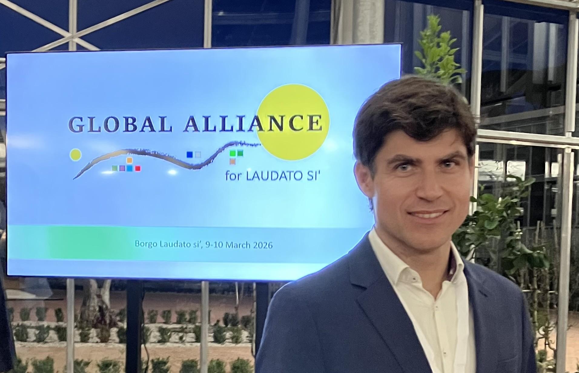 Un hombre sonriente posa frente a un cartel que dice 'GLOBAL ALLIANCE for LAUDATO SI'.